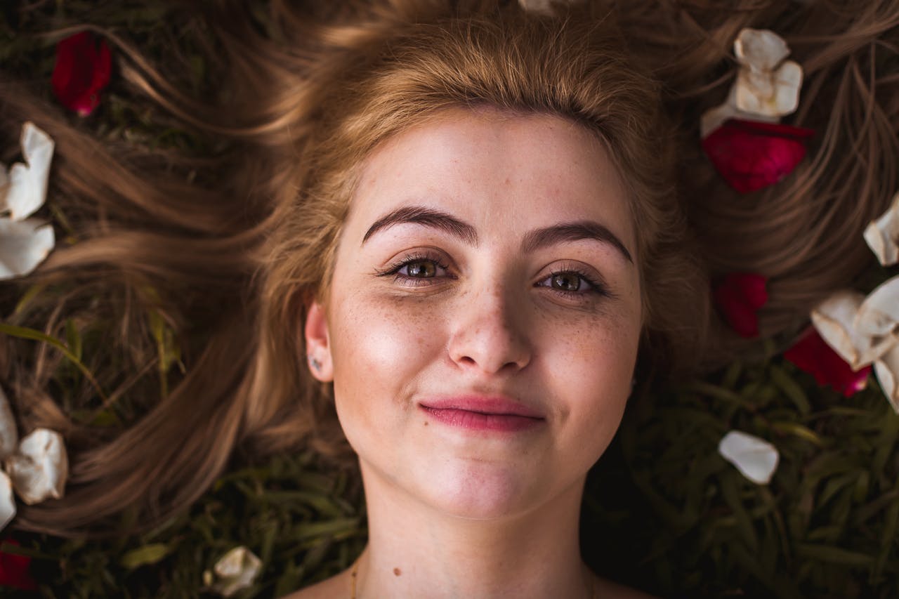 our-services-01 Portrait of a smiling young woman with rose petals in her hair, lying on grass outdoors.